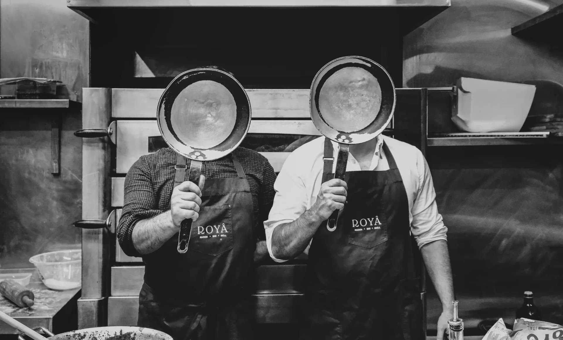Two Royâ chefs in the kitchen, aprons on, pans up